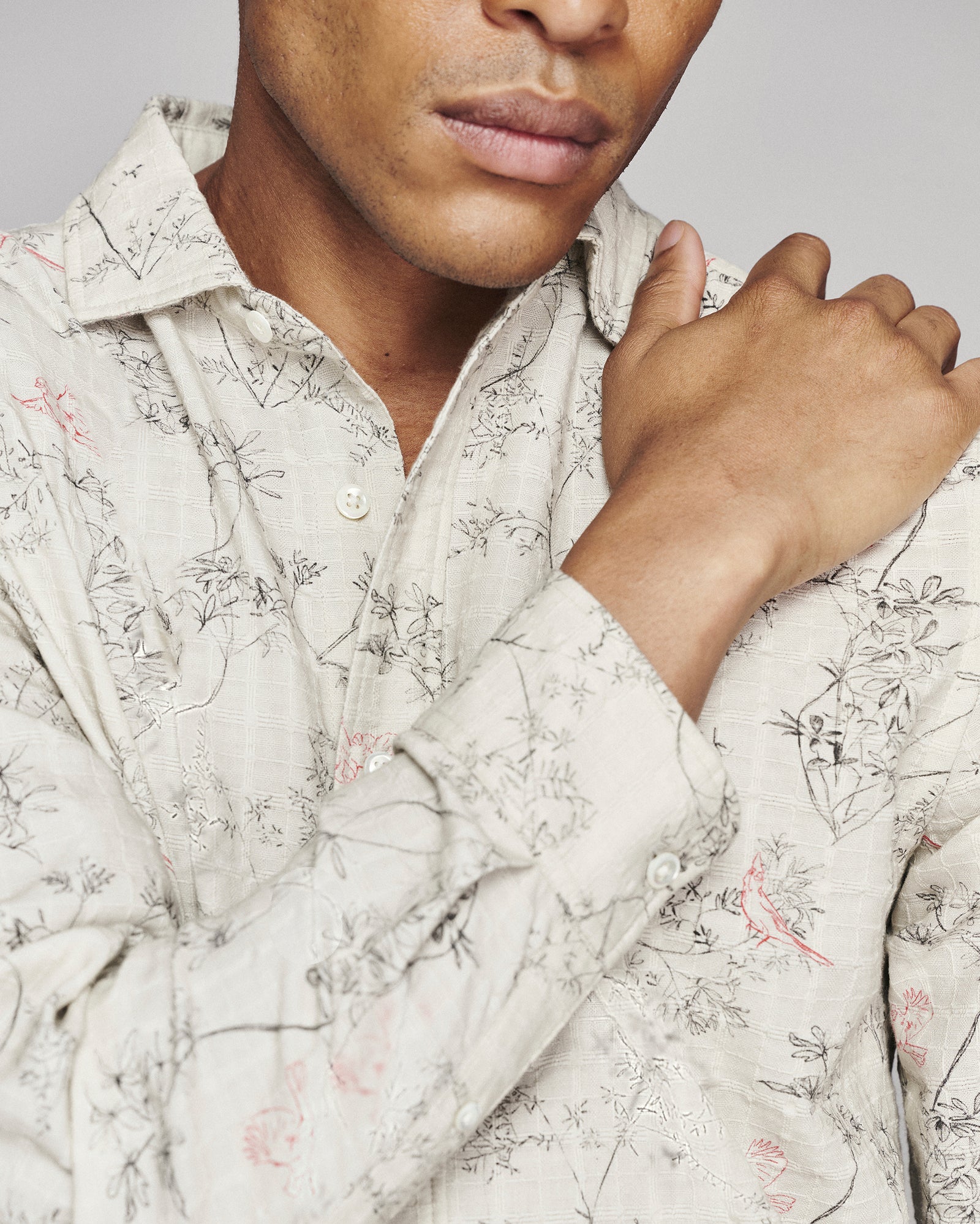 A close-up of a man wearing a light beige long-sleeve button-up shirt with a delicate black and pink botanical pattern, adjusting the collar with one hand.