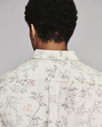Back view of a person wearing a light cream-colored linen shirt with a delicate black botanical print and small pink bird motifs, set against a solid gray background.