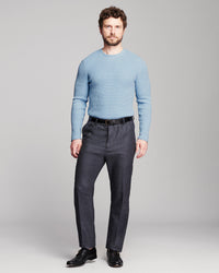 A man modeling a light blue crewneck knit sweater and dark charcoal gray trousers with a black belt and black dress shoes, standing against a neutral gray studio background.
