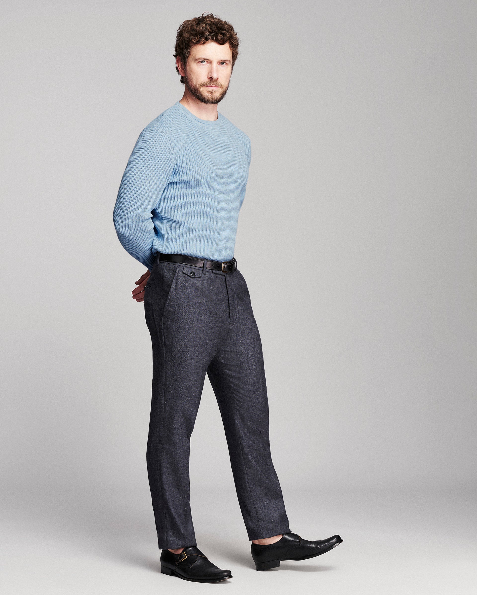 A man modeling a light blue crewneck sweater and dark charcoal gray trousers with black leather dress shoes, standing against a light gray studio background.