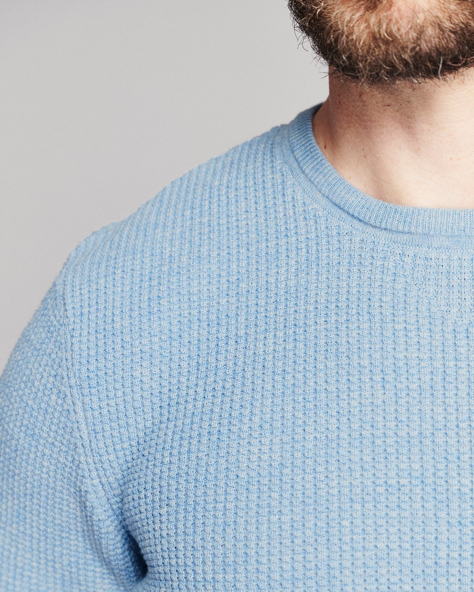 Close-up of a man wearing a light blue textured knit crewneck sweater, showing the ribbed collar and fine waffle-knit fabric against a neutral gray background.