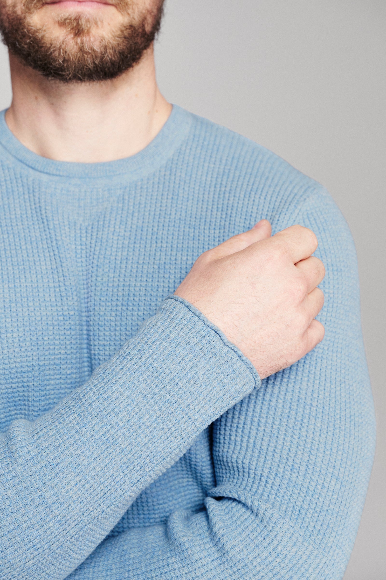 A close-up of a man wearing a light blue waffle-knit crewneck sweater, showing the texture of the fabric and the ribbed cuff.