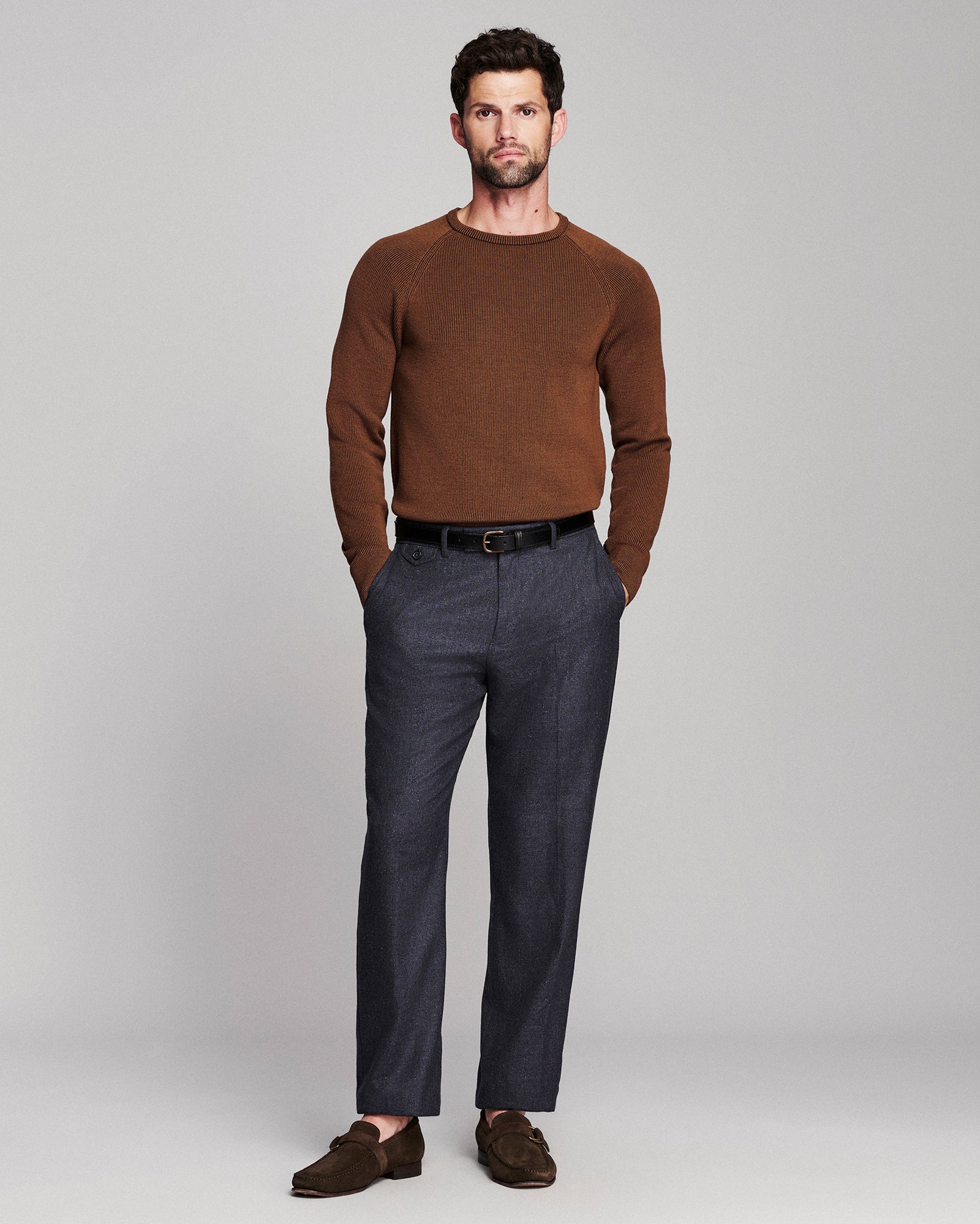 A man modeling a brown crewneck knit sweater, dark gray trousers, a black belt, and brown suede loafers against a light gray studio background.