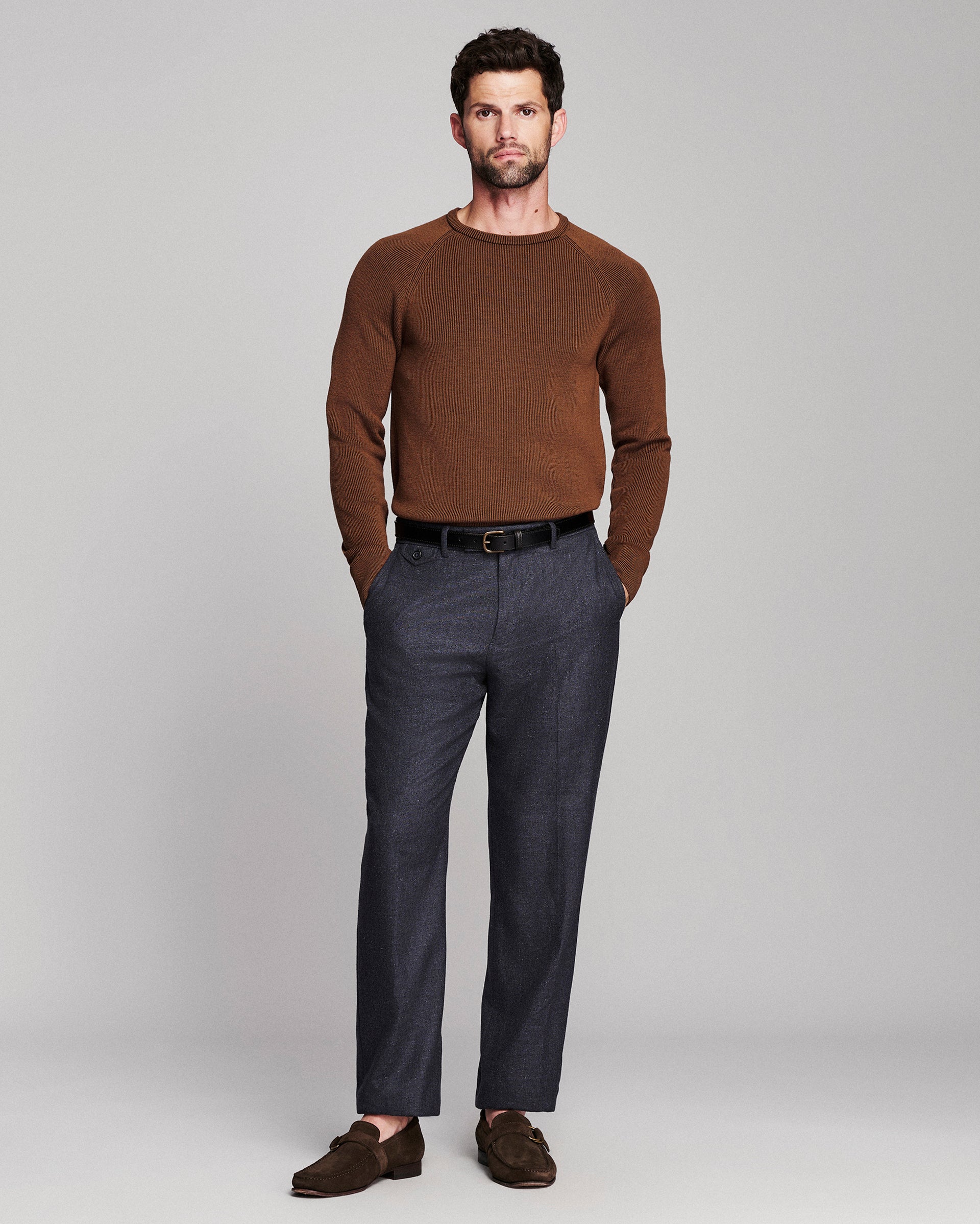 A man modeling a brown crewneck knit sweater, dark gray trousers, a black belt, and brown suede loafers against a light gray studio background.