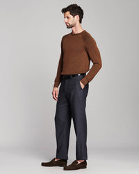 A man wearing a brown long-sleeve knit sweater, dark charcoal blue trousers, a black belt, and dark brown loafers, standing in profile against a light gray studio background.