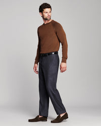 A man modeling a brown knit crewneck sweater, dark blue trousers, a black belt, and brown suede loafers against a light gray studio background.