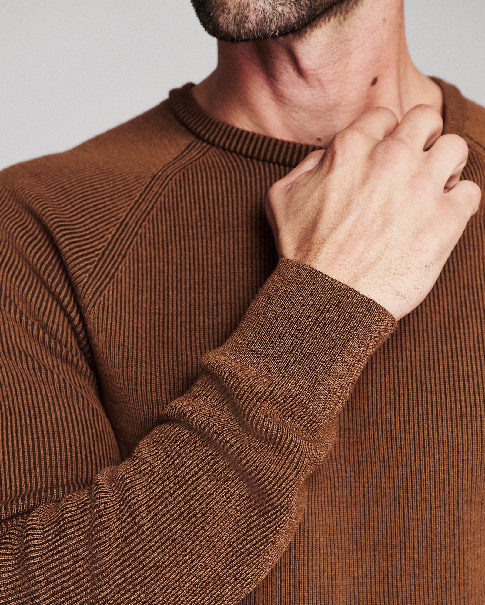 Close-up of a man wearing a brown ribbed knit crewneck sweater, with his hand resting on his neck against a light gray background.