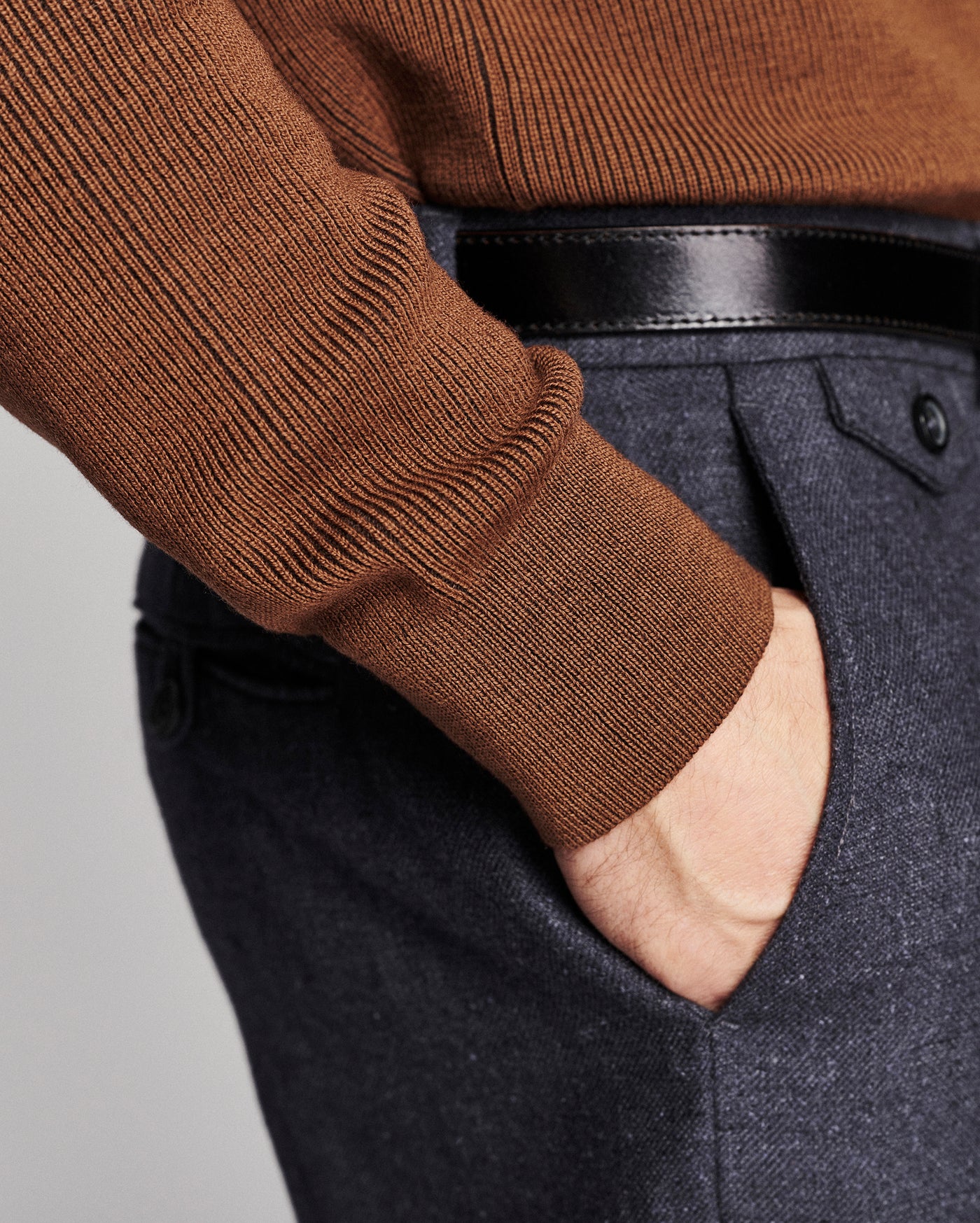 Close-up of a person wearing a brown ribbed knit sweater, a black leather belt, and dark charcoal gray textured trousers with a hand in the pocket.