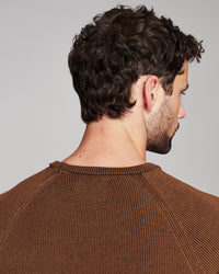 A man shown from the back and side profile wearing a brown ribbed knit crewneck sweater against a plain light gray studio background.