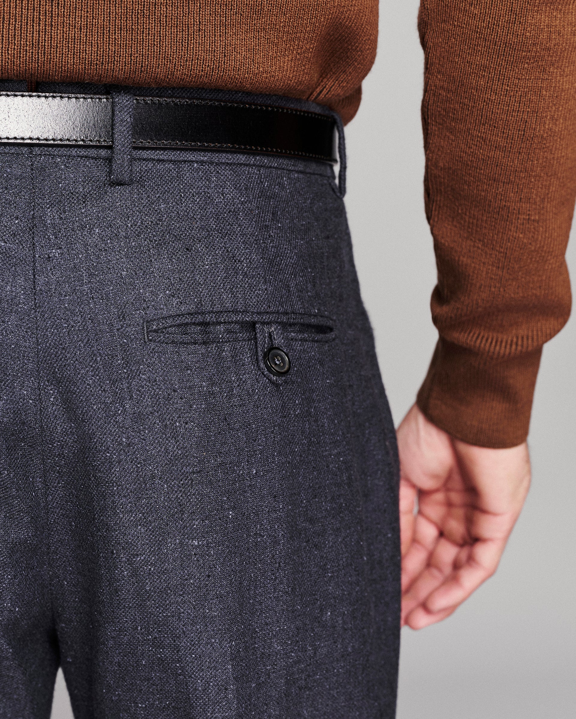 Close-up of a person wearing dark charcoal gray textured trousers with a black belt and a brown knit sweater, showing the back pocket detail.
