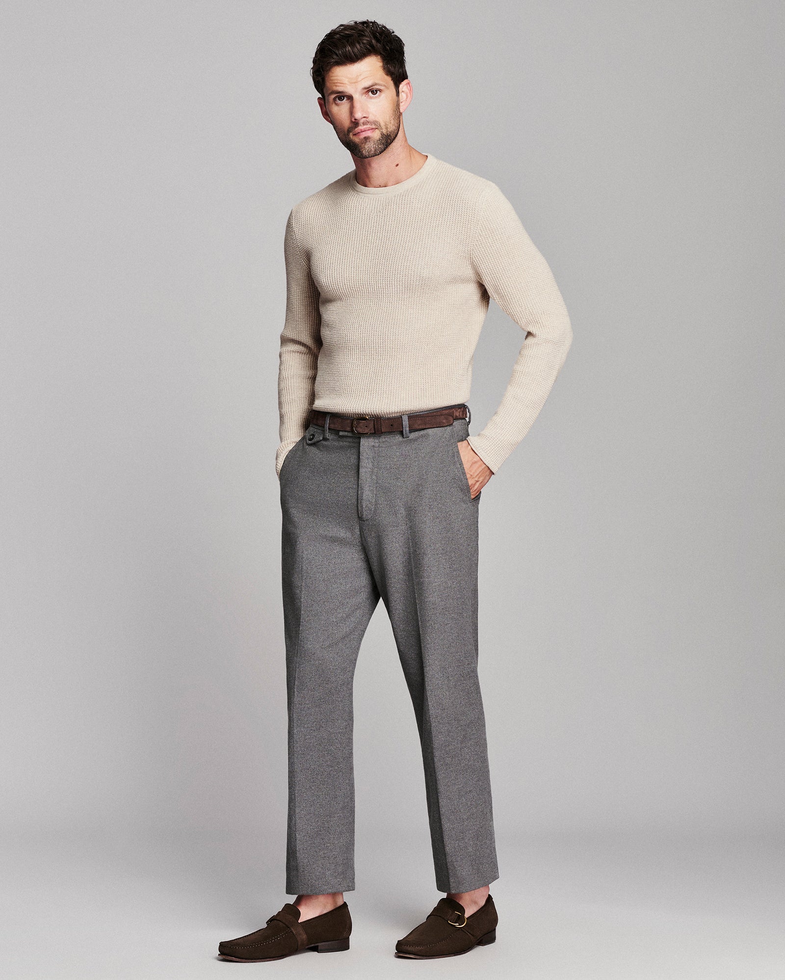 A man modeling a light beige crewneck sweater, gray trousers, a brown belt, and dark brown loafers against a solid light gray studio background.