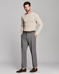 A man modeling a light beige crewneck sweater, gray trousers, a brown belt, and dark brown loafers against a solid light gray studio background.
