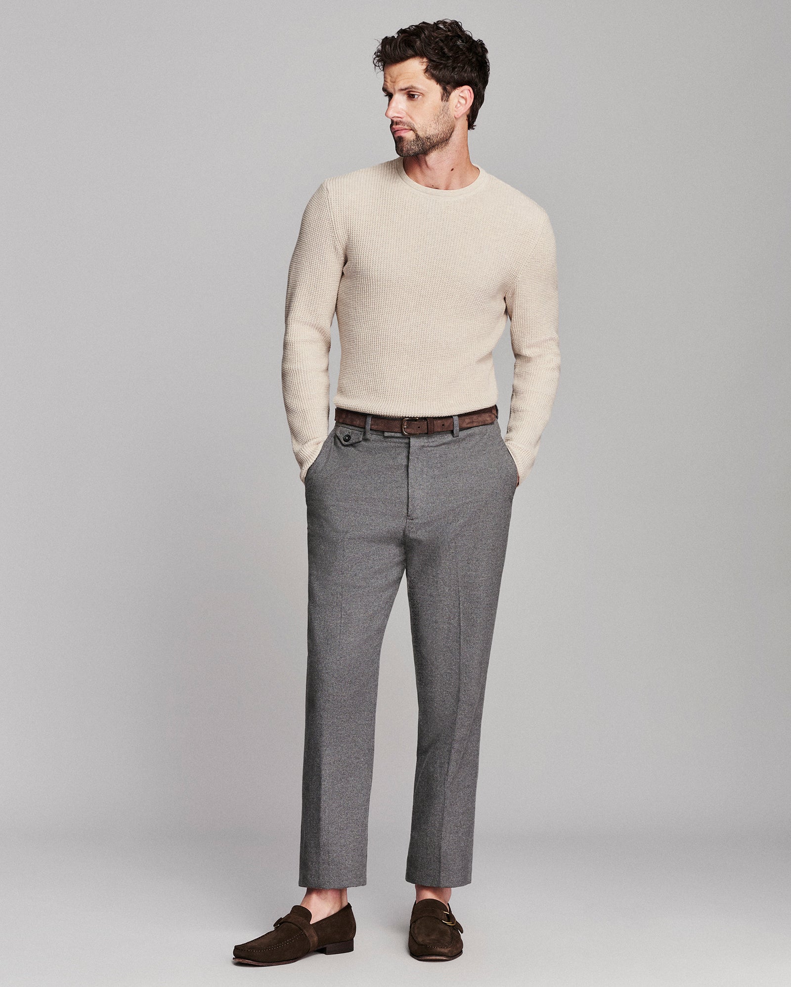 A man modeling a light beige knit crewneck sweater, gray trousers, a brown belt, and dark brown loafers against a solid light gray studio background.
