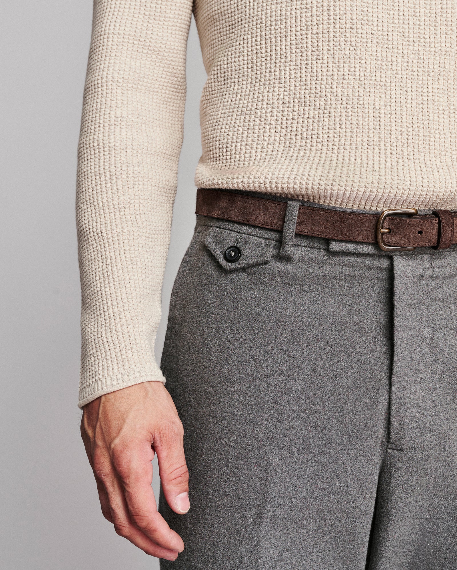 Close-up of a person wearing gray wool trousers, a brown belt, and a cream waffle-knit sweater against a light gray studio background.