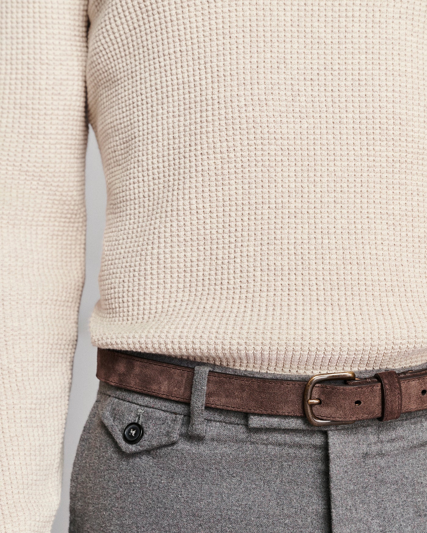 Close-up of a person wearing a cream waffle-knit sweater, a brown leather belt with a brass buckle, and gray wool trousers.