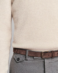 Close-up of a person wearing a cream waffle-knit sweater, a brown leather belt with a brass buckle, and gray wool trousers.