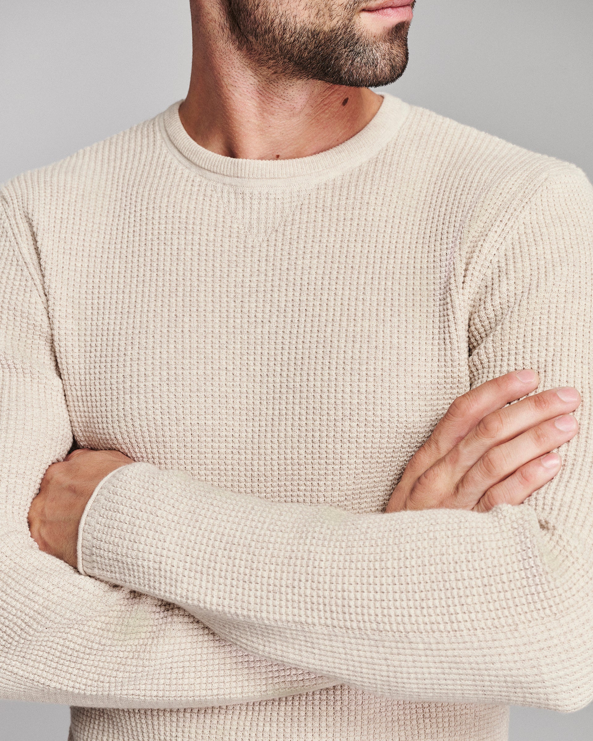 A close-up of a man wearing a textured cream-colored knit sweater with his arms crossed, set against a plain gray studio background.