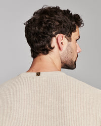 A man shown from the back and side profile wearing a cream-colored textured knit sweater against a solid gray background.