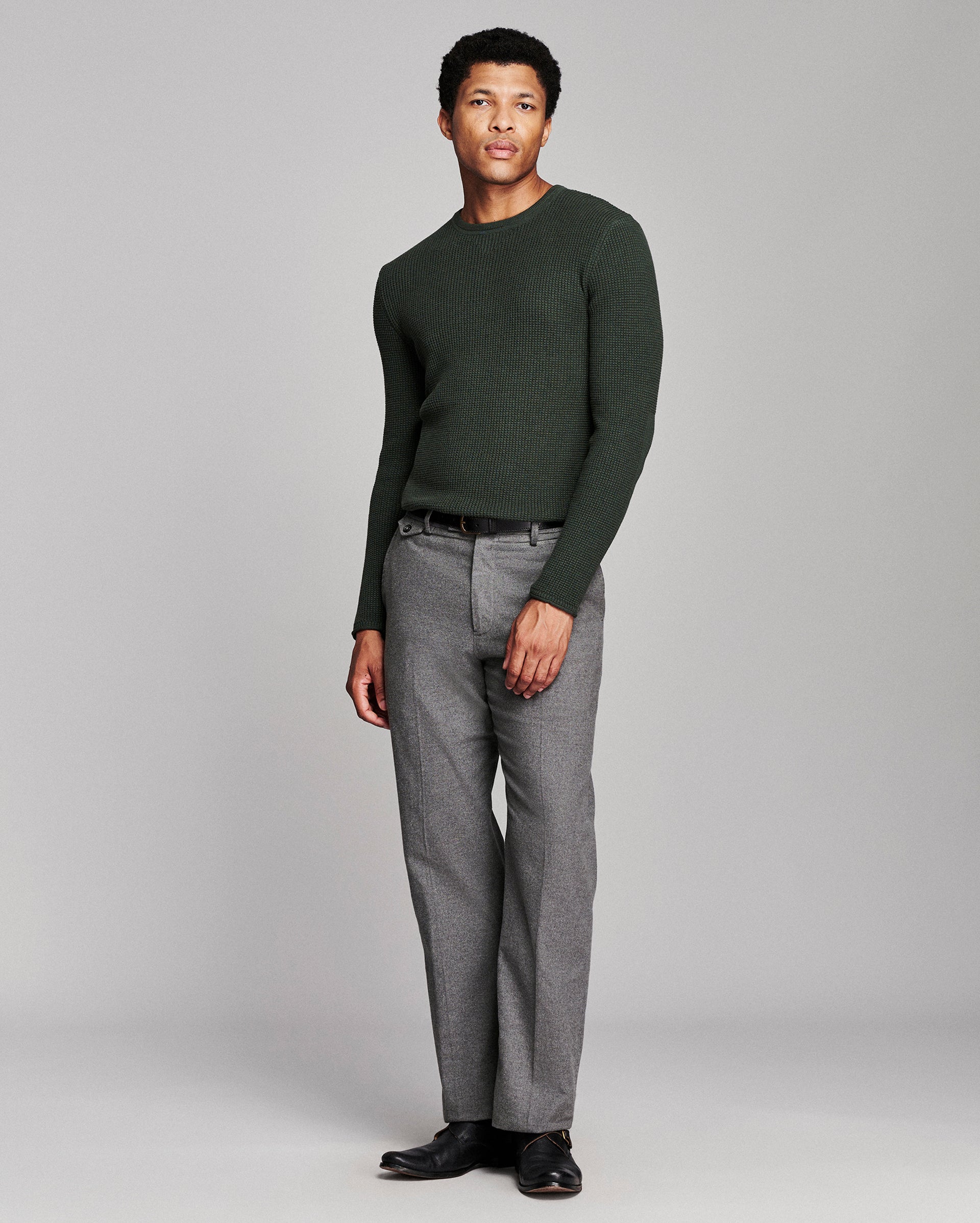 A man modeling a dark forest green long-sleeve knit sweater and gray trousers against a light gray studio background.
