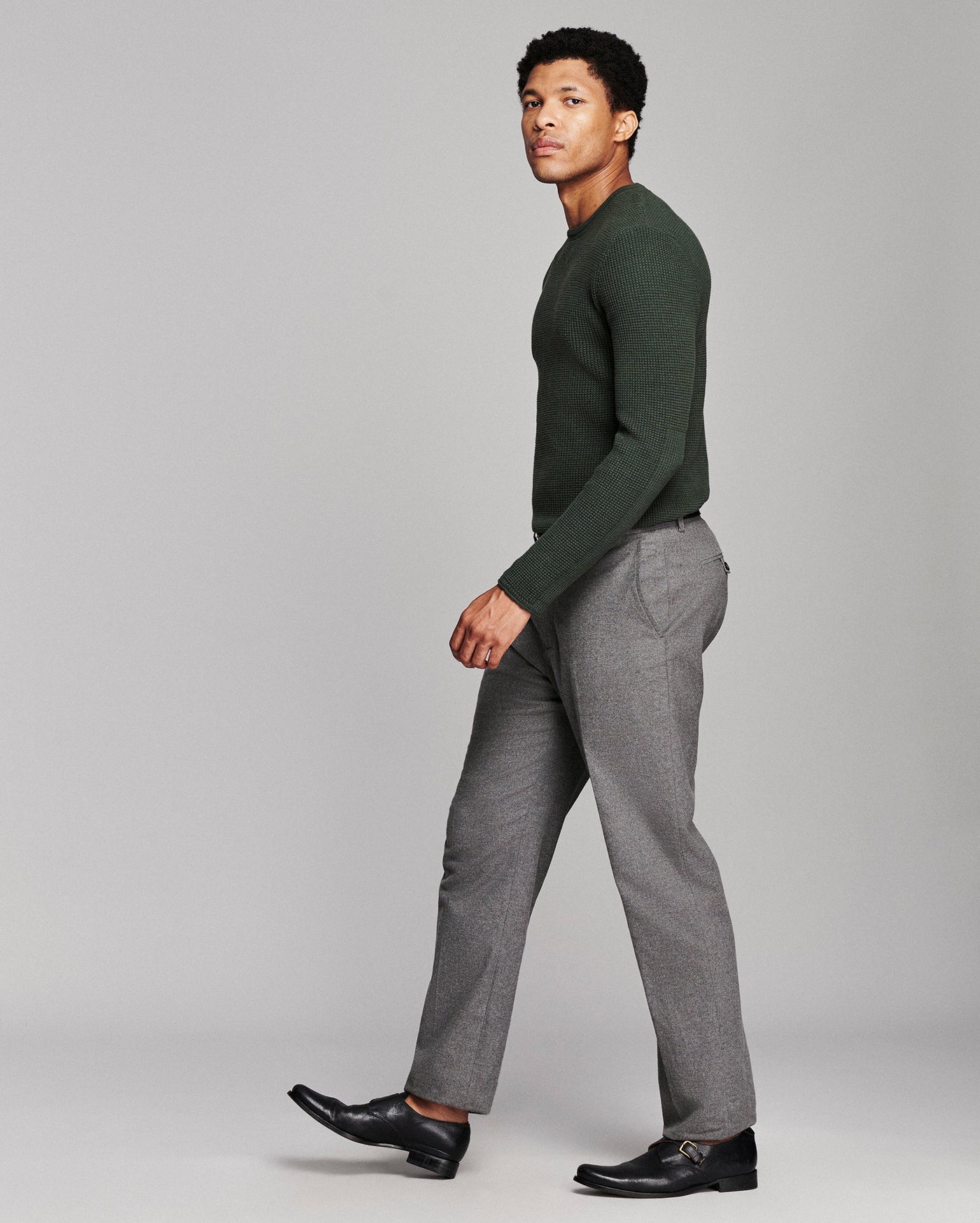 A man in a dark green long-sleeve knit sweater and gray trousers walking in profile against a light gray studio background.