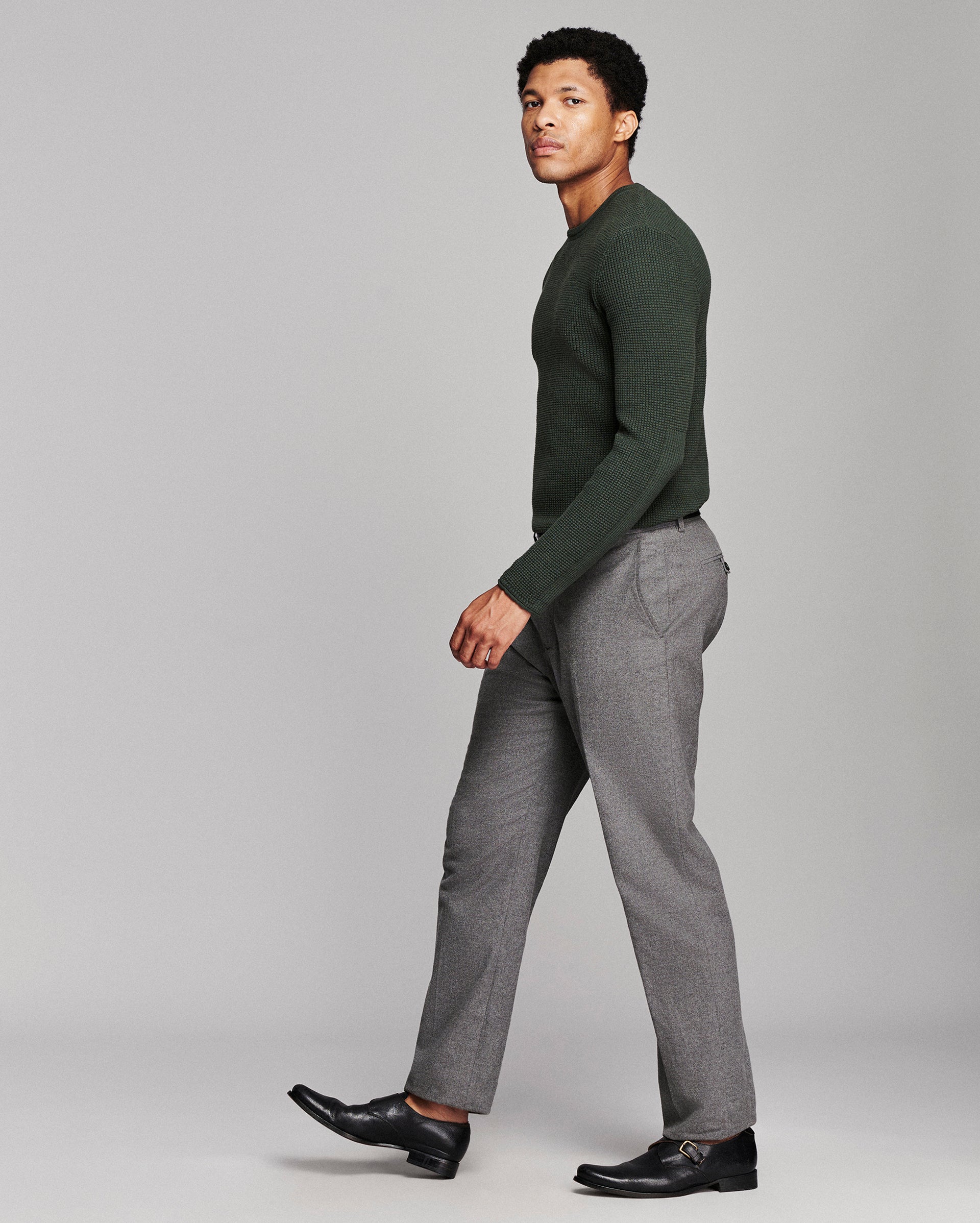 A man in a dark green long-sleeve knit sweater and gray trousers walking in profile against a light gray studio background.