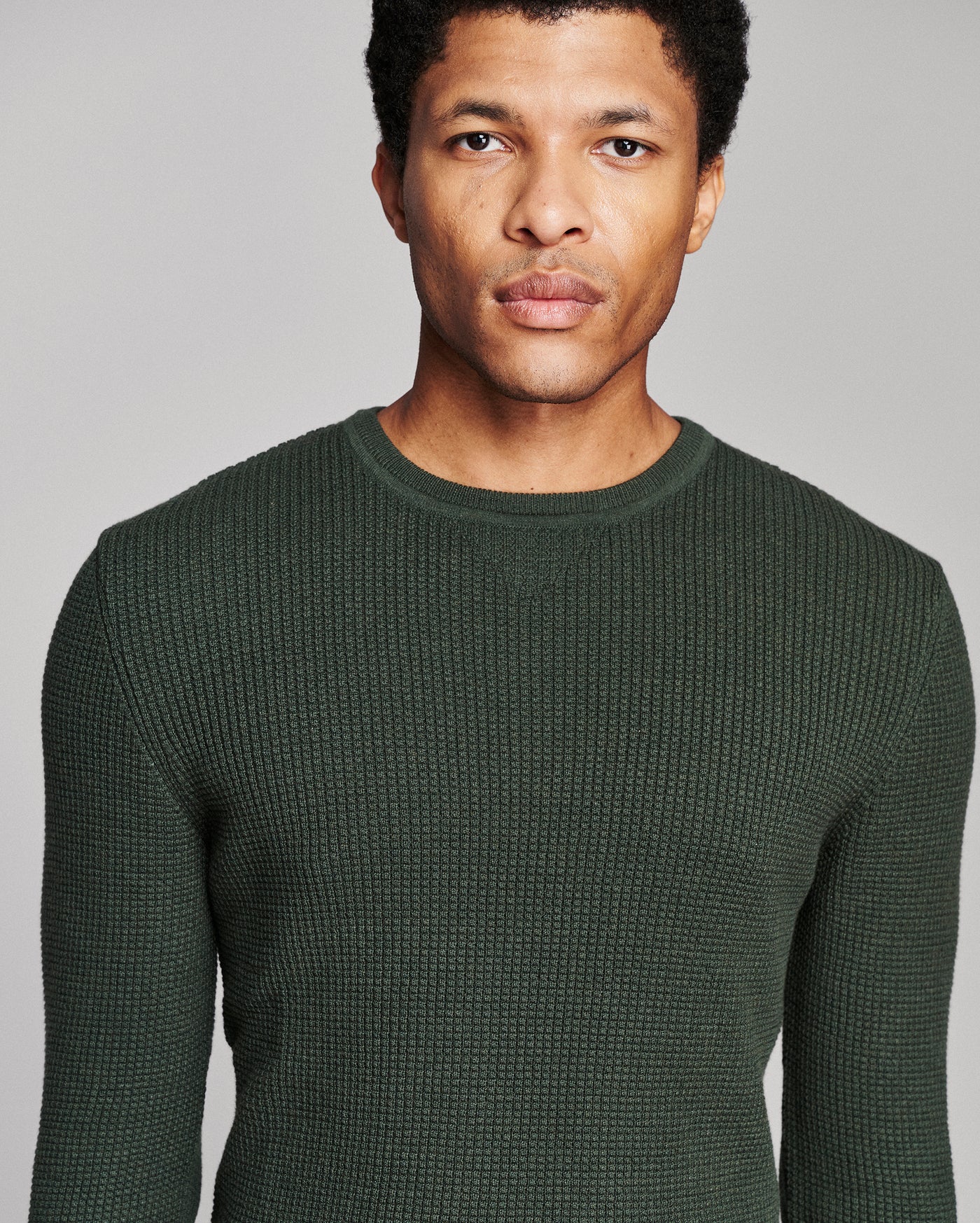 A man wearing a dark forest green textured knit crewneck sweater, shown from the chest up against a solid light gray background.