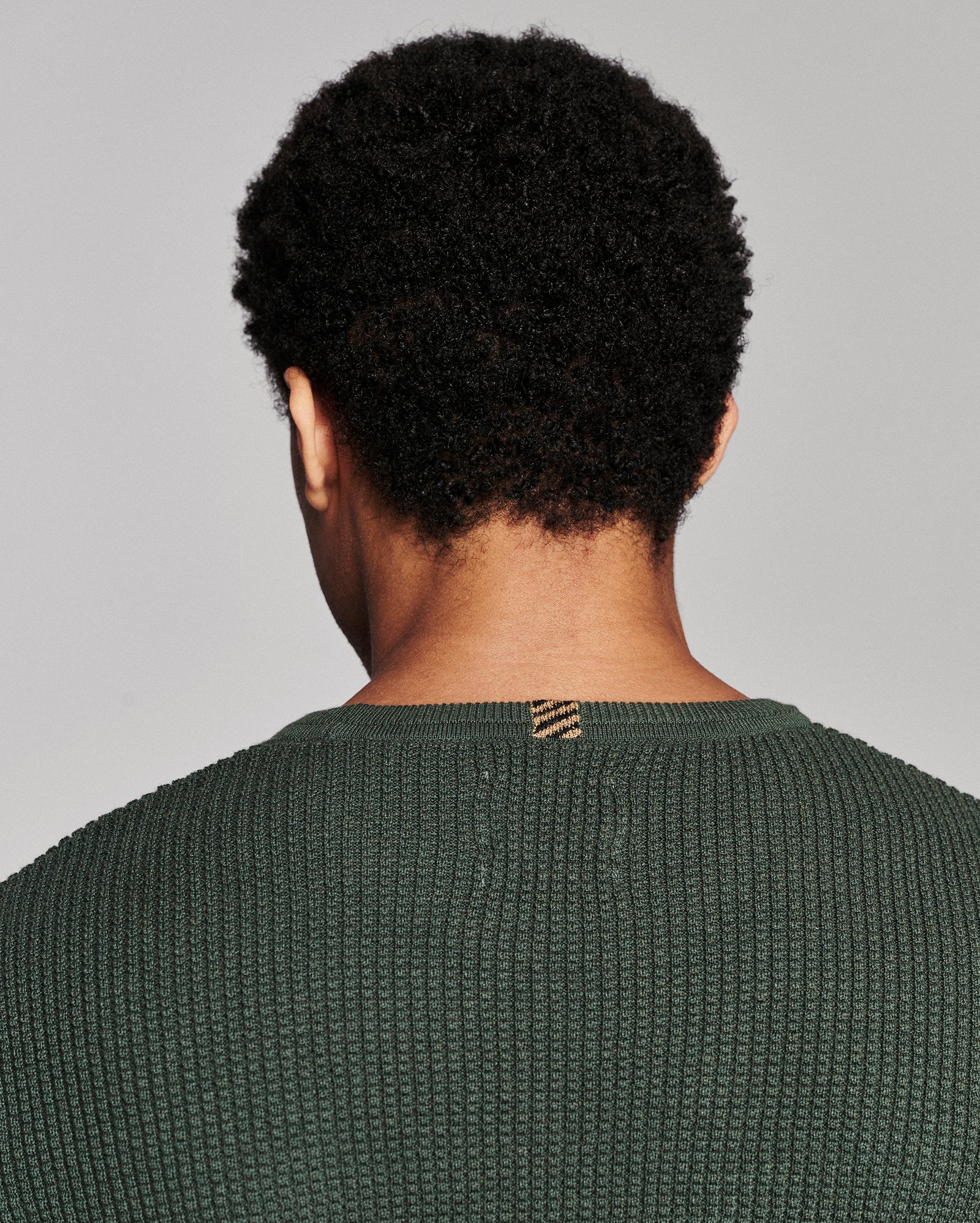 Back view of a person with short curly black hair wearing a textured dark green knit sweater against a light gray studio background.