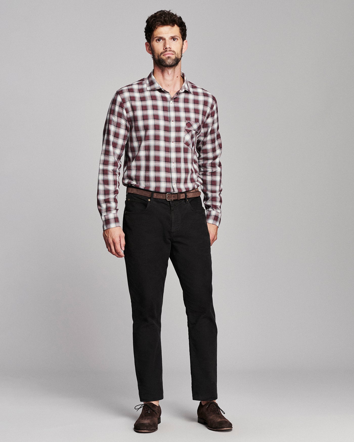 A man modeling a long-sleeve button-down plaid shirt in red, white, and black, paired with black trousers and brown leather shoes, against a solid light gray background.