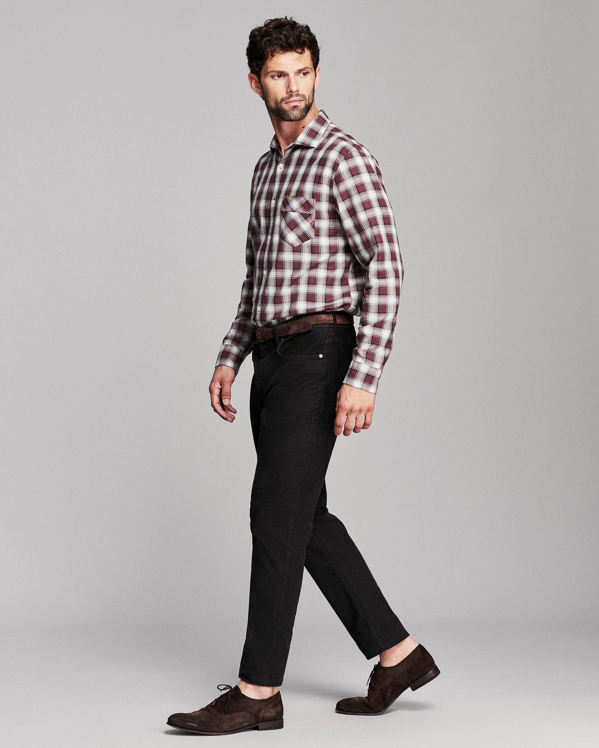 A man modeling a long-sleeve red and white plaid button-down shirt, paired with black jeans, a dark brown belt, and dark brown suede oxford shoes, against a solid light gray background.