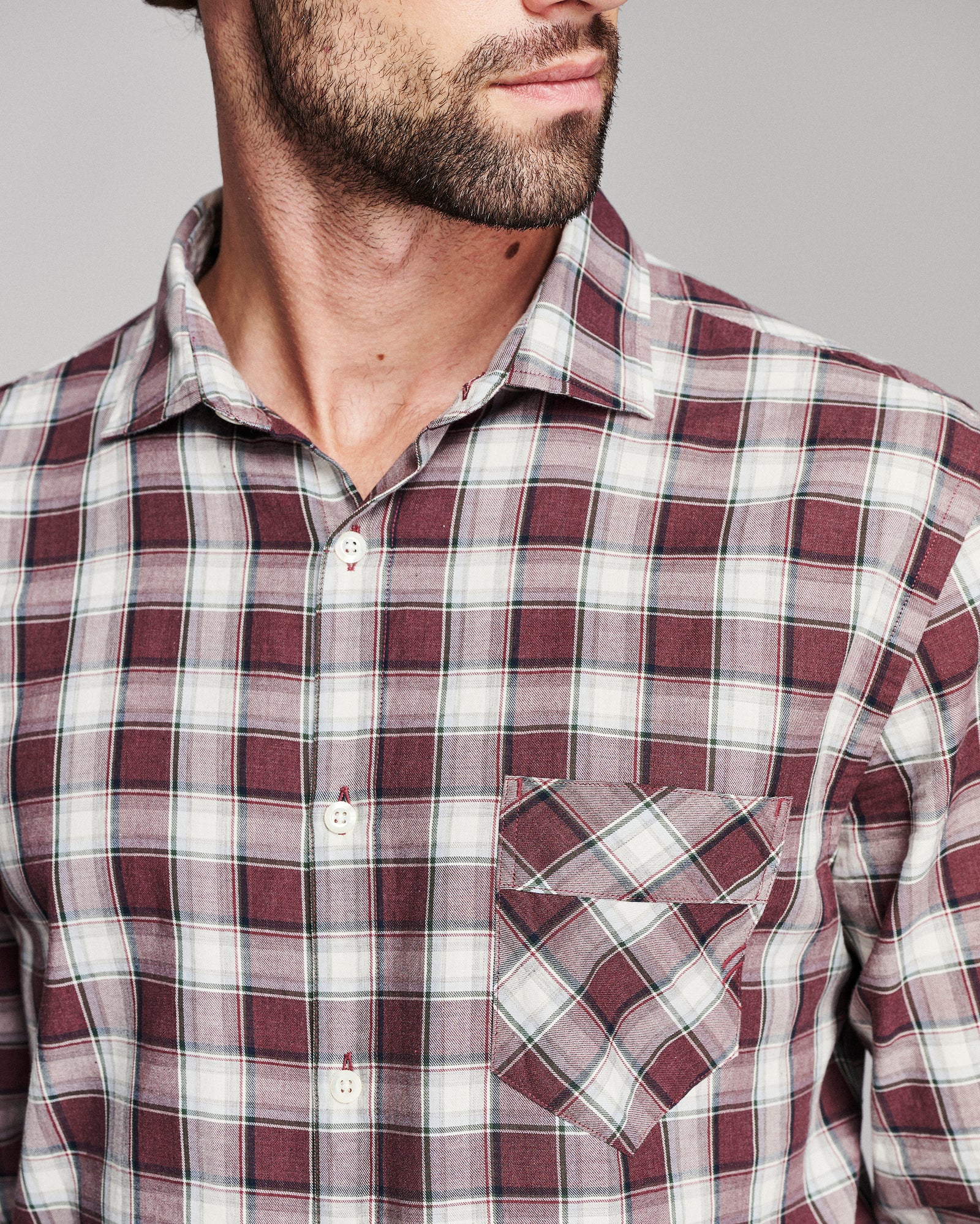 A close-up of a man wearing a burgundy and white plaid button-down shirt with a chest pocket, shown from the chin down against a neutral gray background.