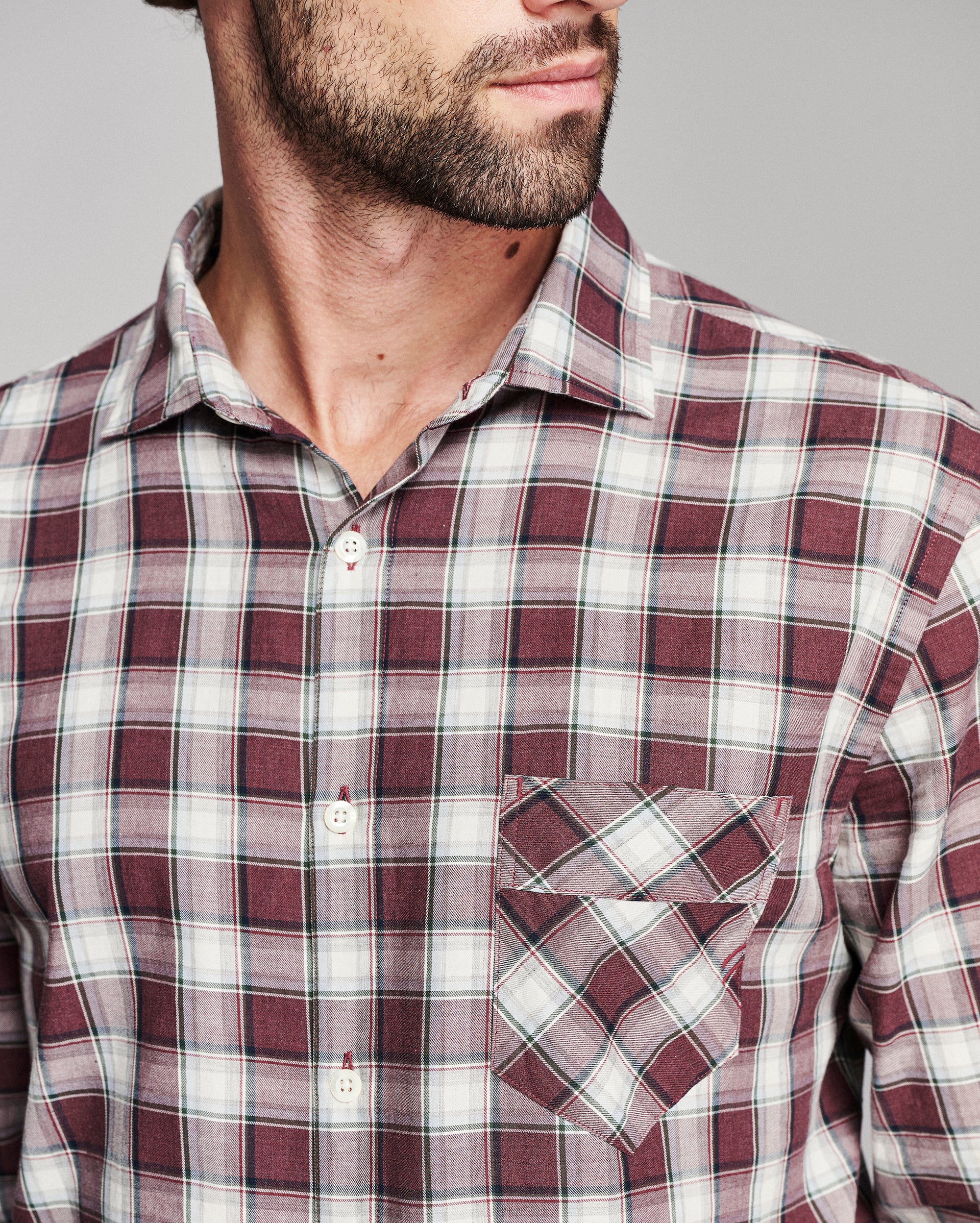 A close-up of a man wearing a burgundy and white plaid button-down shirt with a chest pocket, shown from the chin down against a neutral gray background.