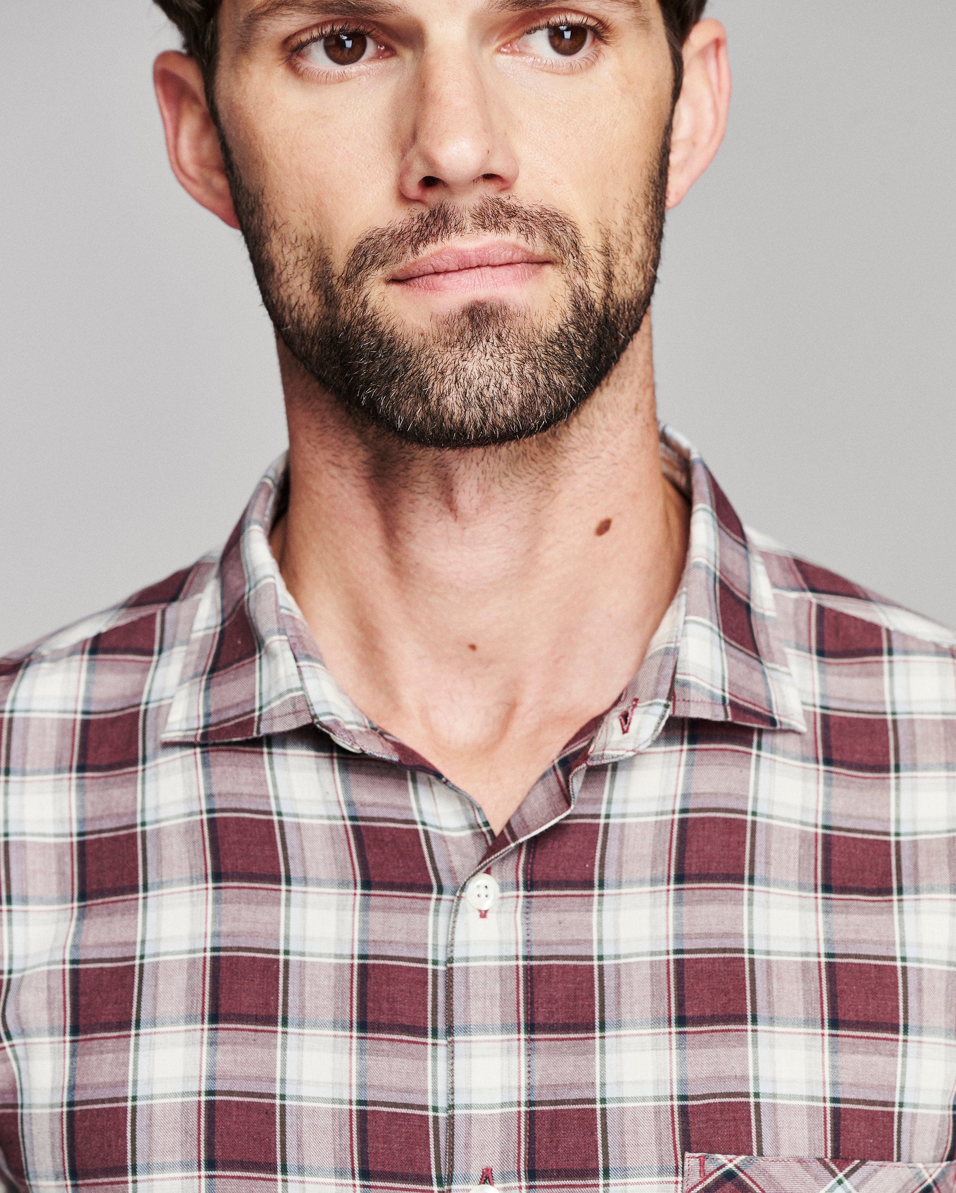 A close-up shot of a man wearing a maroon, white, and gray plaid button-down shirt against a solid gray background.