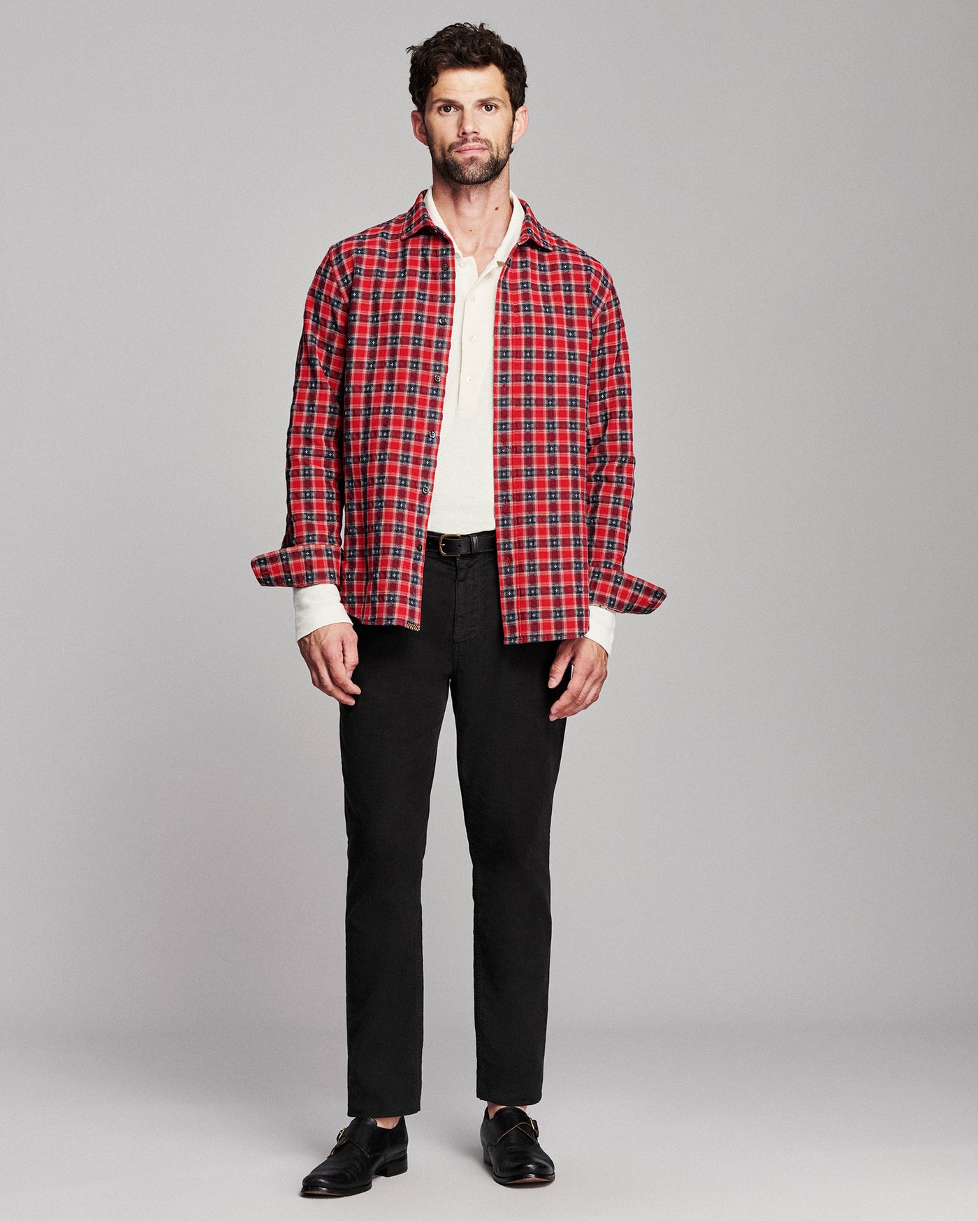 A man modeling a red and black plaid button-down shirt over a white collared shirt, paired with black trousers and black shoes, against a solid gray studio background.