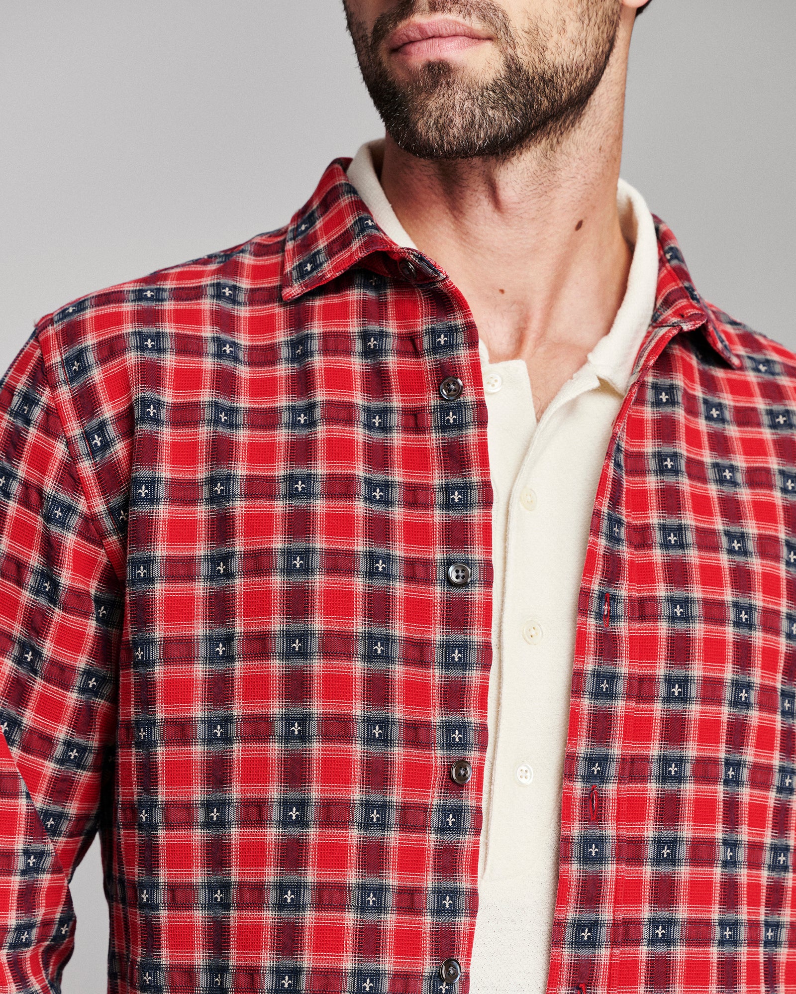 A close-up of a man wearing a red and navy plaid button-down shirt over a cream collared undershirt, against a plain gray studio background.