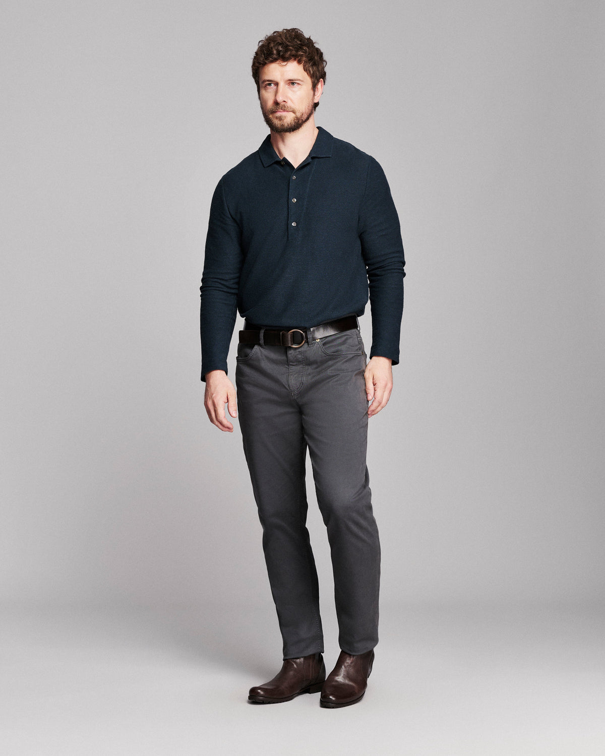 A man modeling a dark navy long-sleeve polo shirt, dark gray dress pants, a black belt, and brown leather shoes against a solid light gray studio background.