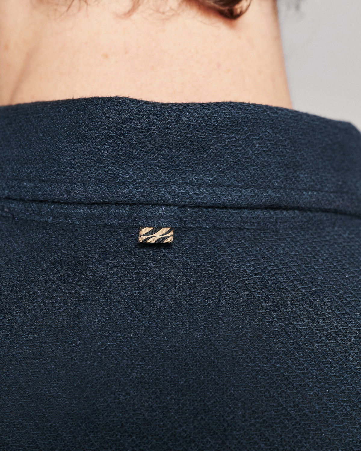 Close-up of a person's neck and upper back wearing a dark navy textured knit top, featuring a small gold-tone striped metal brand pin on the collar.