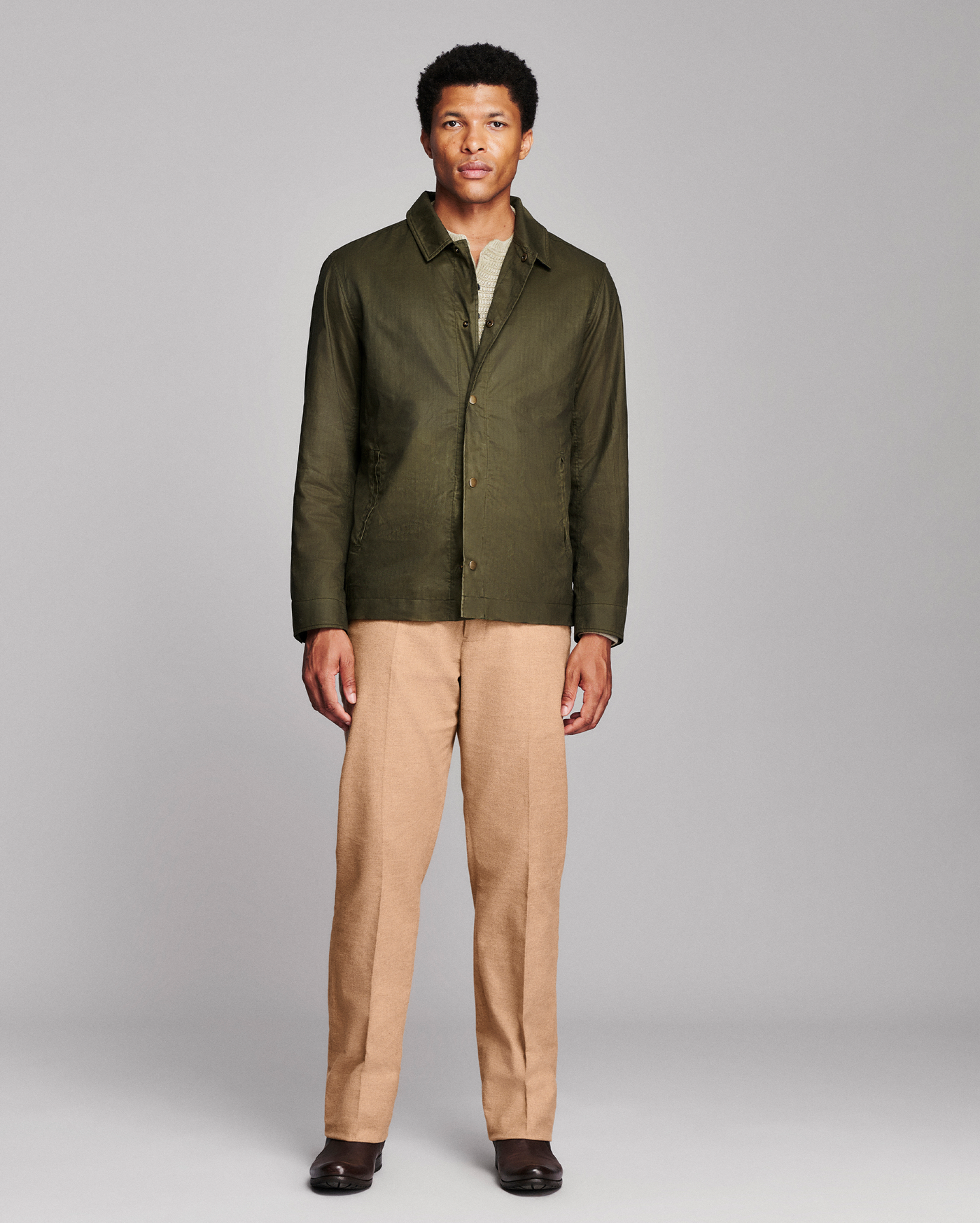 A man modeling an olive green button-up shirt jacket over a light-colored undershirt, paired with tan trousers and dark brown shoes, against a solid gray studio background.
