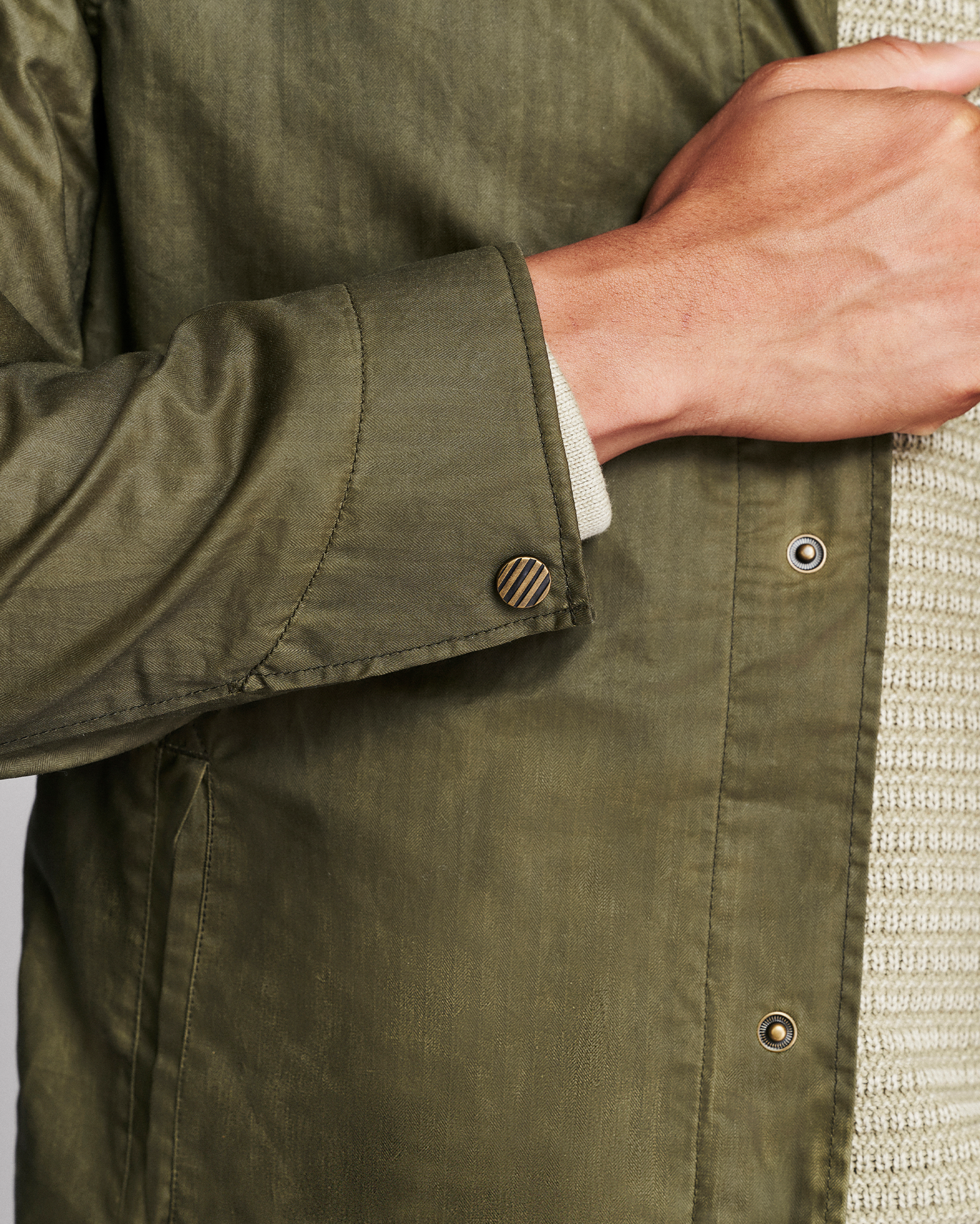 Close-up of an olive green button-up shirt worn by a model, showing the buttoned cuff, fabric texture, and a ribbed knit sweater underneath.