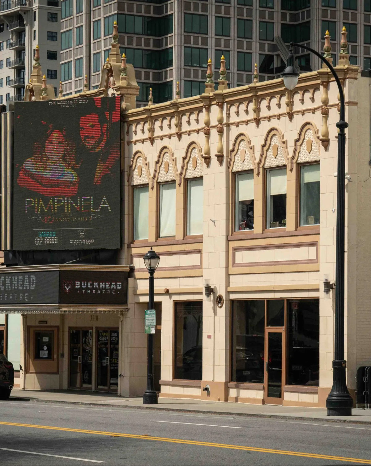 A city street scene showing a beige ornate building facade with storefront windows, a large dark movie poster reading PIMPINELA on the left, and a Buckhead Theatre sign below. Tall modern buildings are visible in the background behind the structure.