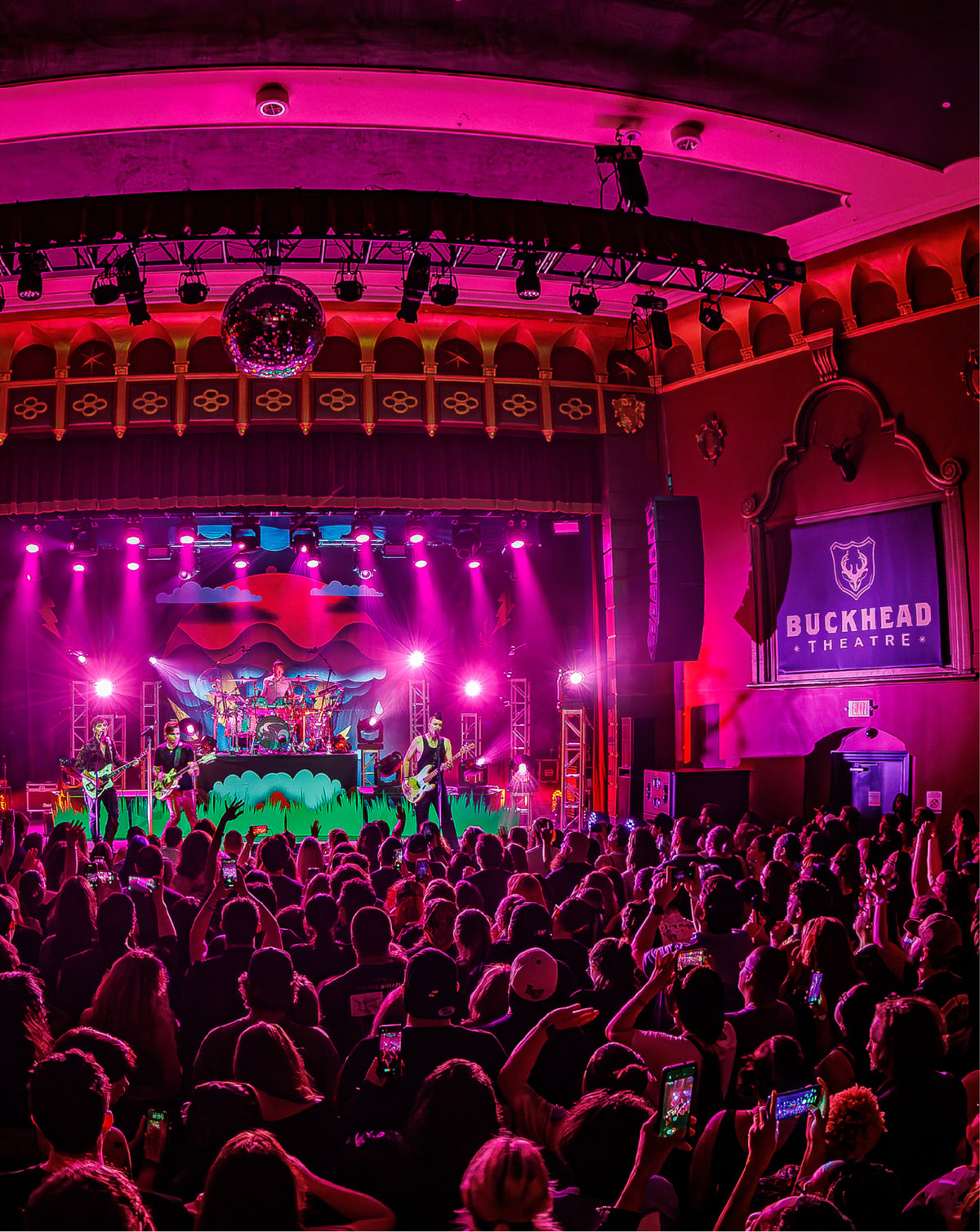 A live concert performance at Buckhead Theatre, with a band on stage under pink and purple stage lights, a large audience in the foreground, and a screen displaying the Buckhead Theatre logo.
