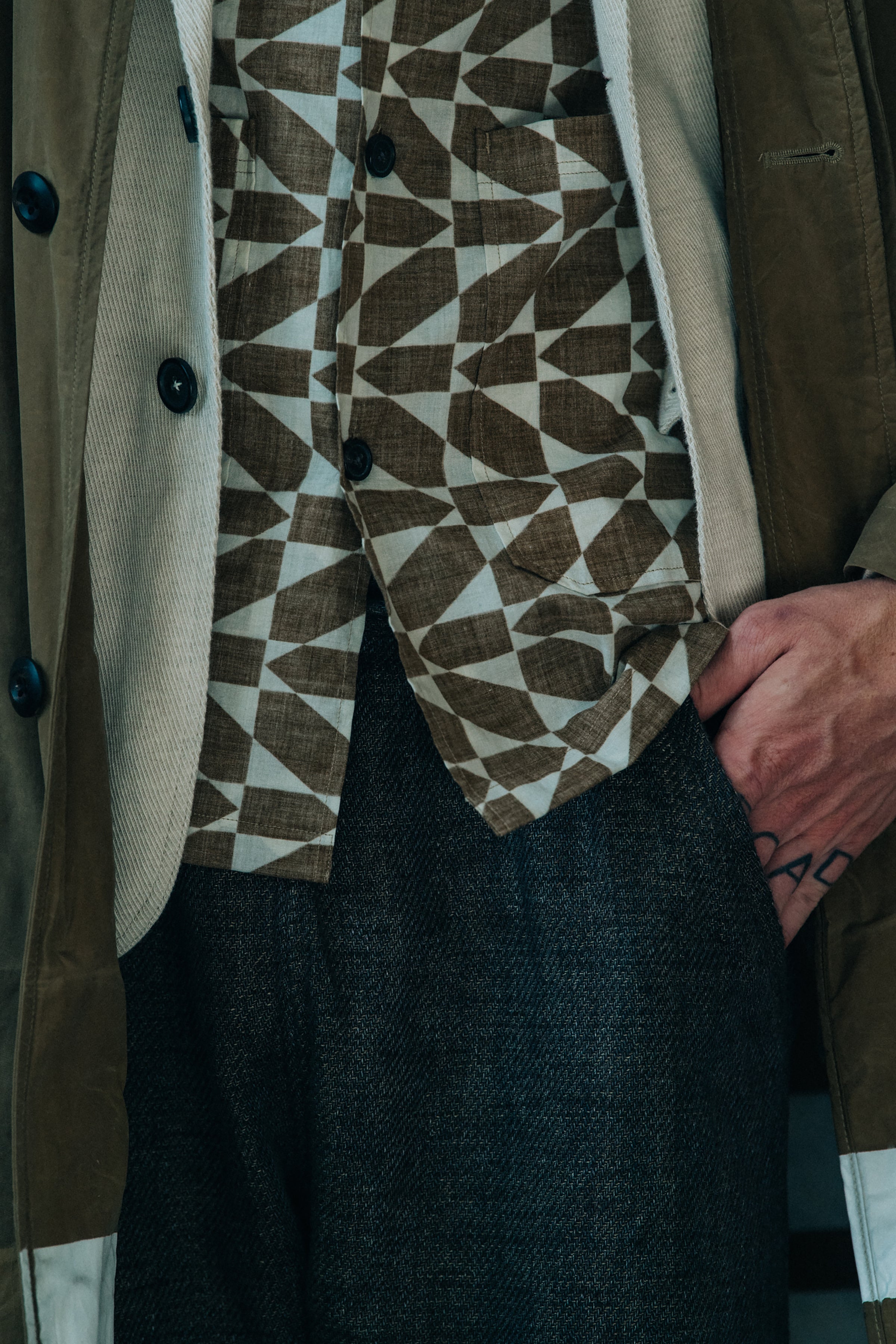 A close-up of a person wearing a brown and beige geometric patterned waistcoat over a cream shirt, paired with a brown jacket and dark gray trousers.