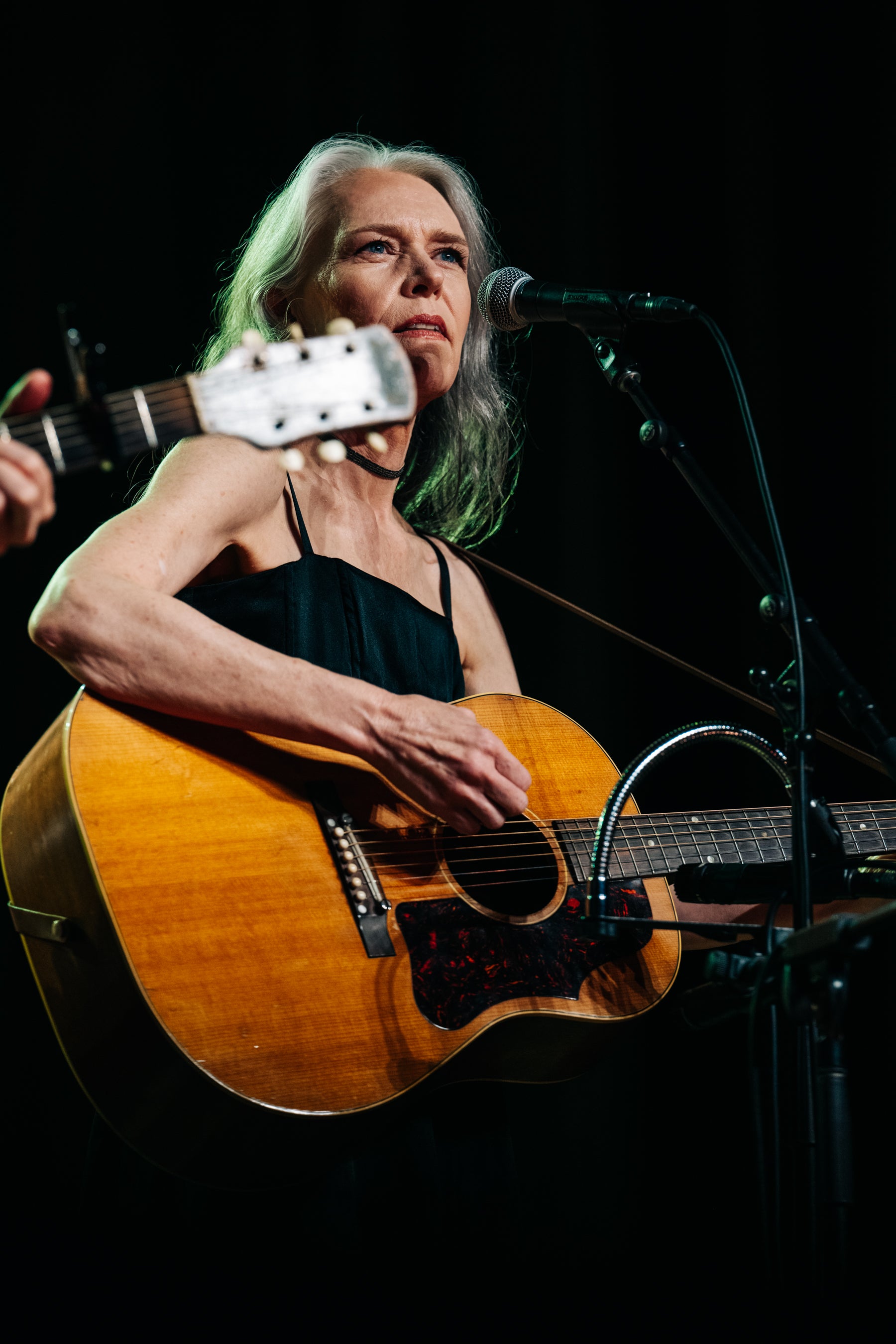 A woman with gray hair playing an acoustic guitar on stage, holding a microphone in front of her, against a dark background.