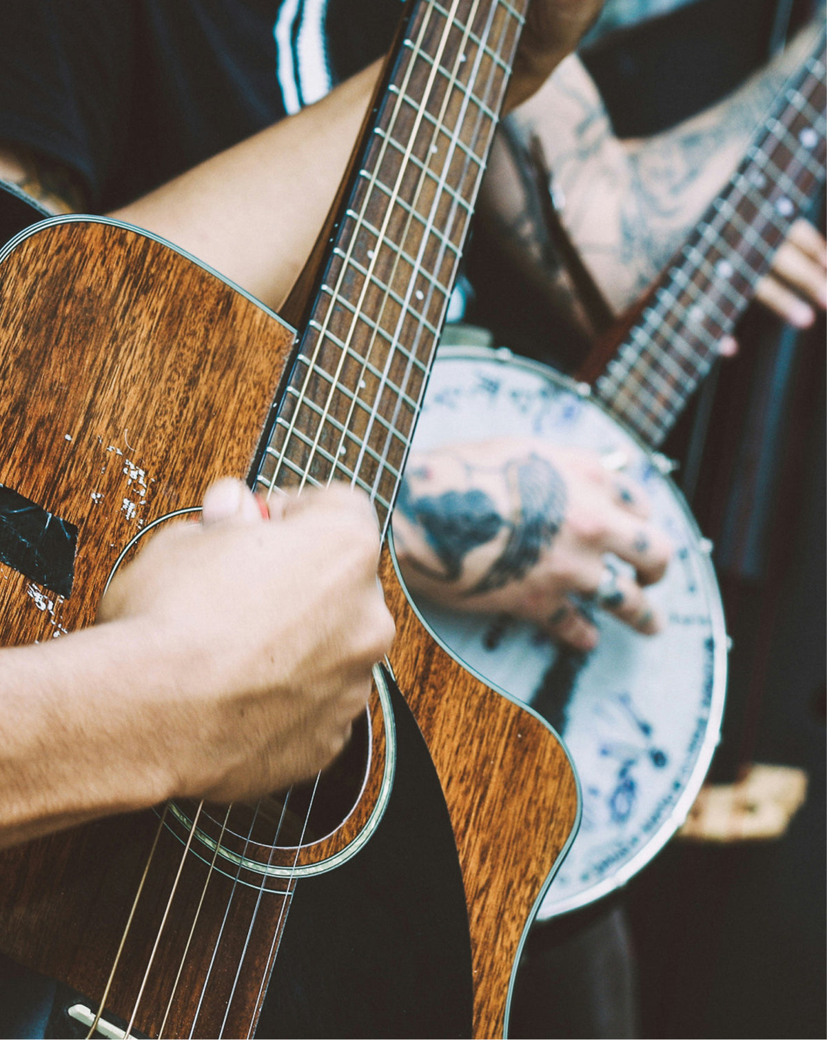 Close-up of a person playing an acoustic guitar, with tattooed hands visible on the neck and strumming, while another person holds a banjo in a dimly lit setting.