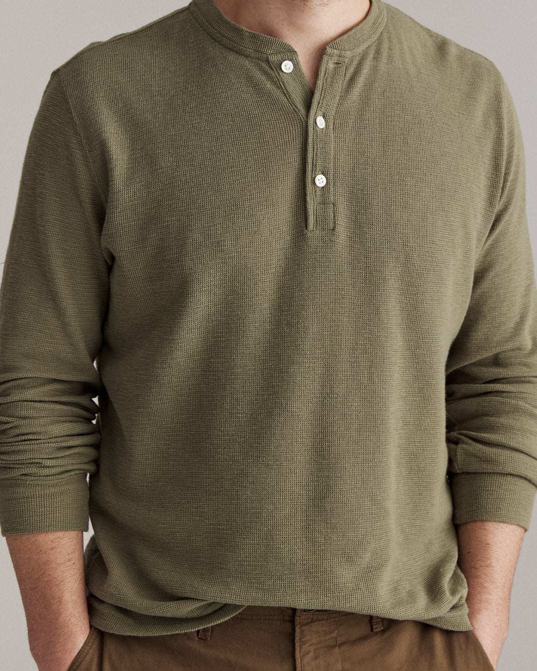 A person wearing an olive green, long-sleeve Henley shirt with three white buttons, and brown pants. The persons face is partially out of frame.