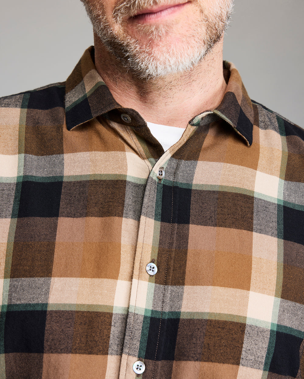 A close-up of a man wearing a brown, black, and beige plaid button-up shirt. His face is partially visible, showing a light beard and a subtle smile. The shirt has a collar and white buttons.