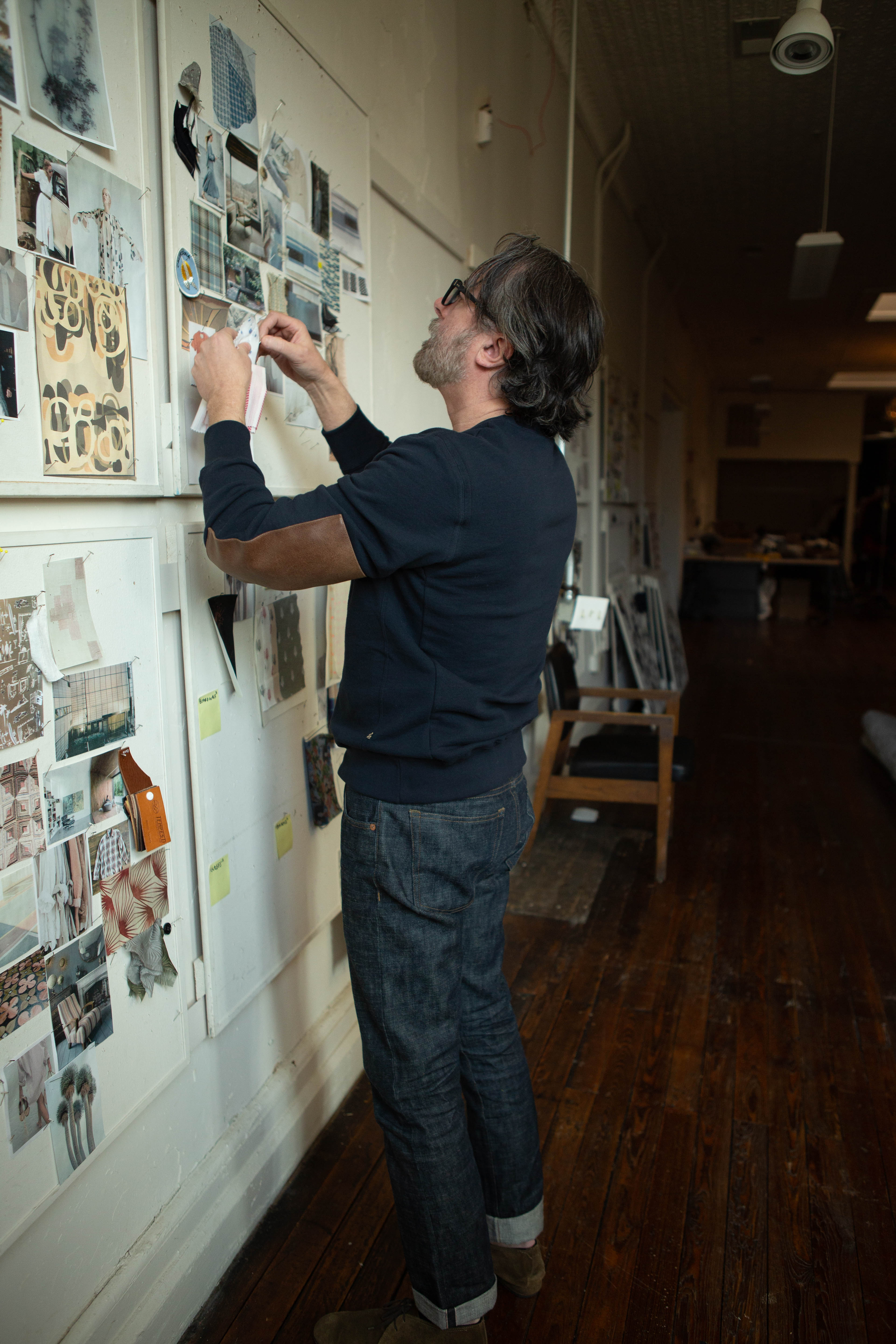 A man wearing a dark navy long-sleeve shirt and blue jeans stands in a hallway, carefully placing a small printed card or sticker onto a white wall covered with photos and notes.