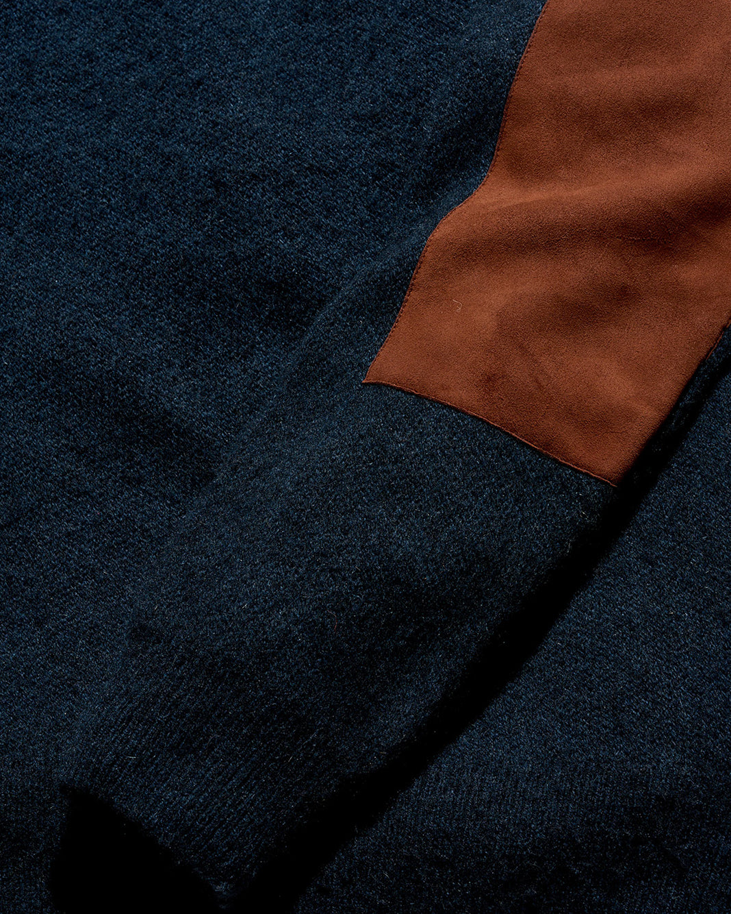 Close-up of a knit sweater showing a dark navy blue body with a contrasting rust-brown suede elbow patches.