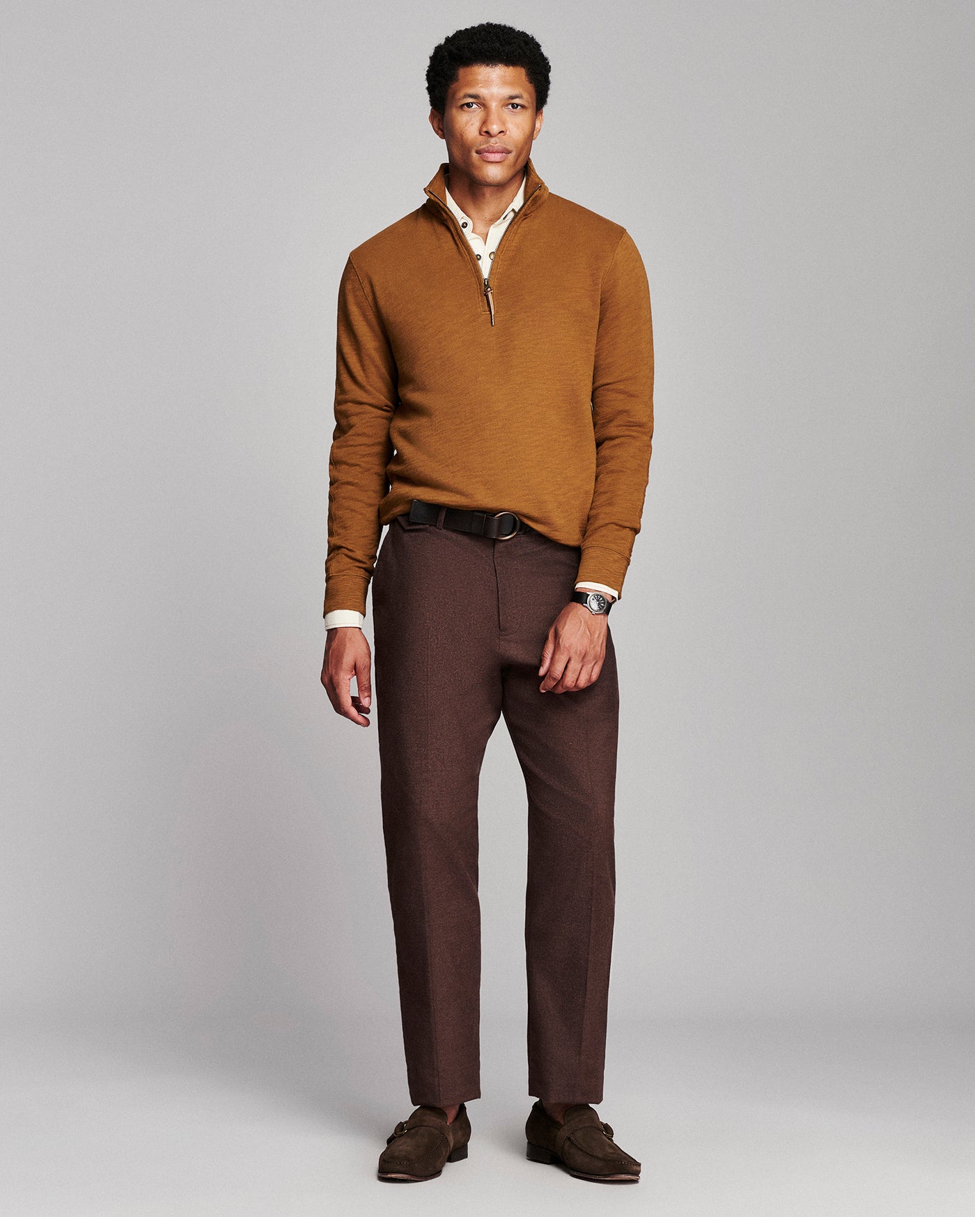 A man modeling a brown quarter-zip knit sweater over a cream collared shirt, paired with dark brown trousers and brown loafers, against a solid light gray studio background.