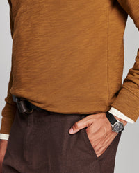 A close-up of a person wearing a textured mustard-brown long-sleeve knit top and dark brown trousers, with one hand in a pocket and a black watch on the wrist.