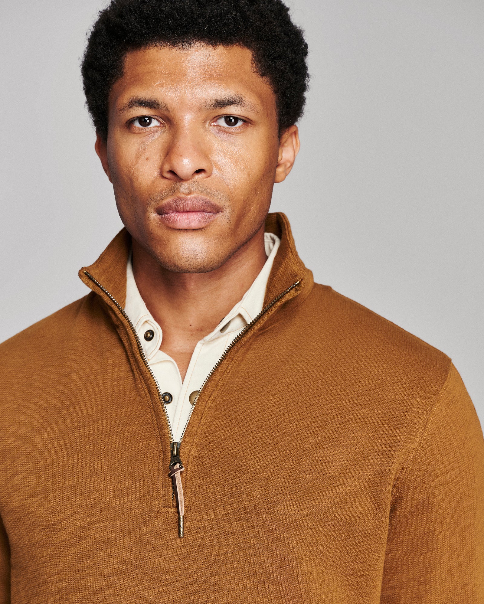 A man wearing a brown quarter-zip pullover sweater over a cream button-down shirt, photographed against a solid light gray background.